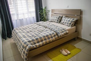 En snyggt bäddad säng med ett rutigt överkast och matchande kuddar, en trägavel, en grön matta på golvet och ett par tofflor på mattan.