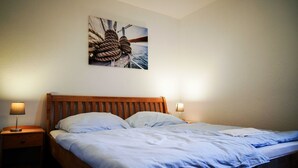 Ein ordentlich bezogenes Bett mit einem hölzernen Kopfteil, zwei Nachttischlampen und einem Bild von einem Schiffsmast und Seilen an der Wand.