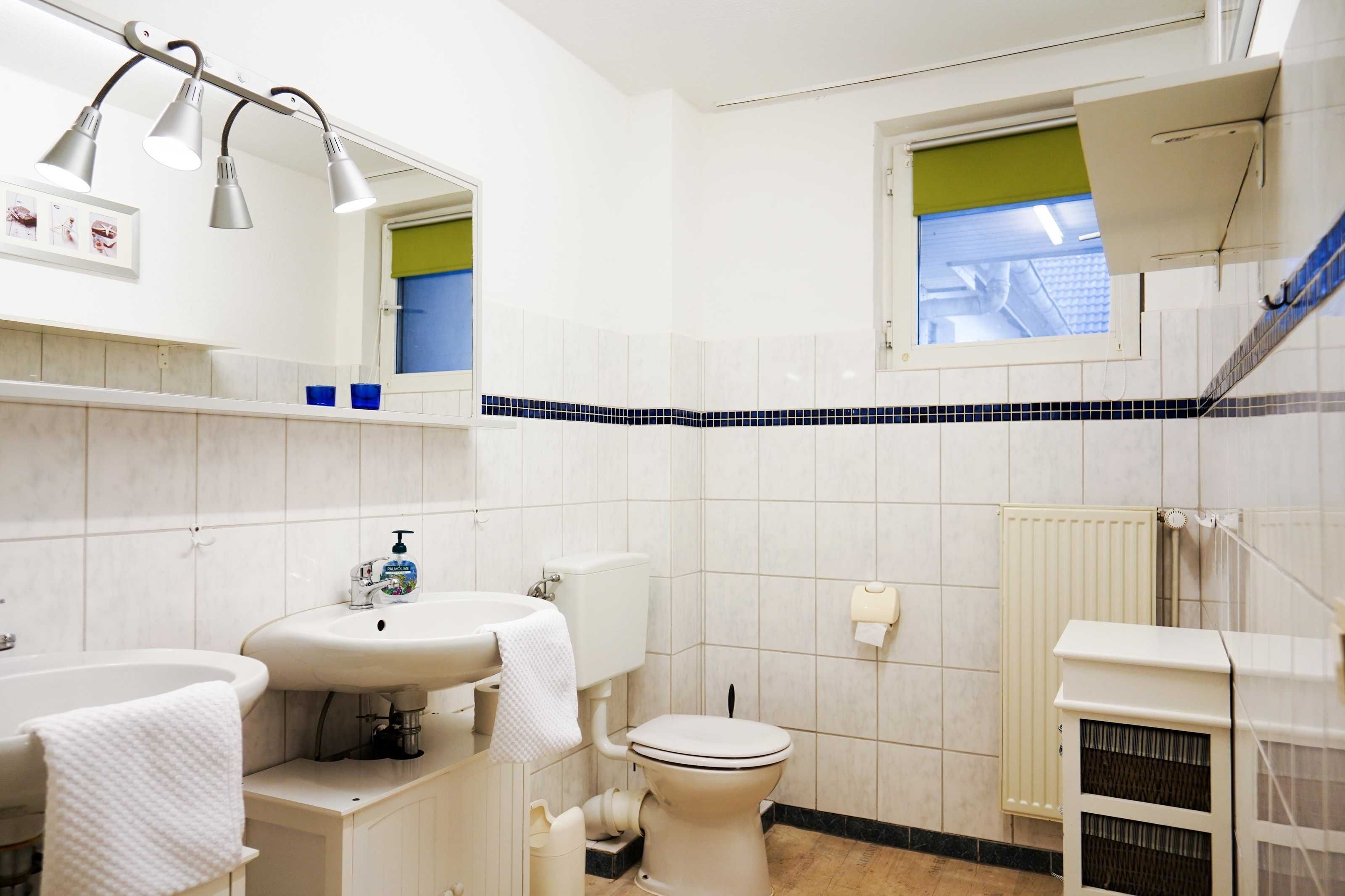 Ein Badezimmer mit einem weißen Waschbecken, einer Toilette und einer Badewanne. Ein Fenster mit grüner Jalousie und ein Heizkörper an der Wand sind vorhanden.
