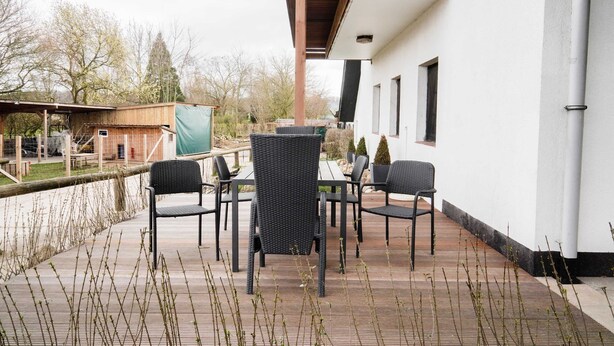 Eine überdachte Terrasse im Freien mit Tisch und Stühlen, eine Holzterrasse und ein Blick auf die Umgebung mit Bäumen und einem Gebäude.