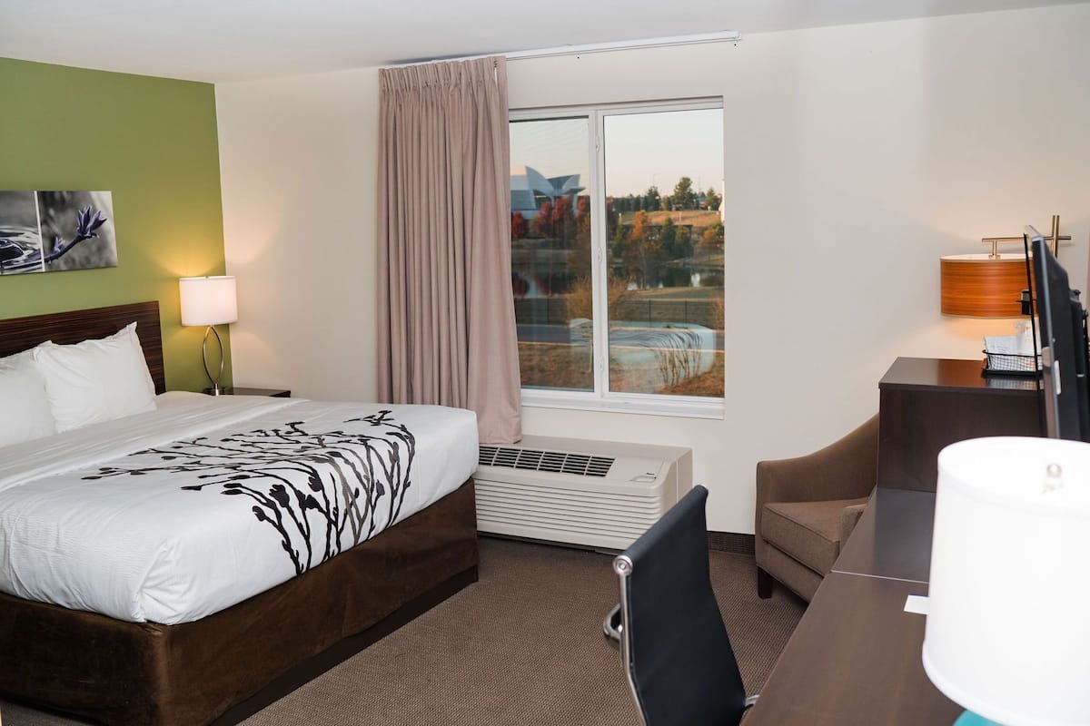 A hotel room with a bed, a desk, a chair, a window with a view, and a wall-mounted air conditioner.