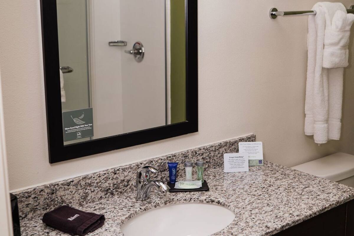 A bathroom with a large mirror, a sink, a towel rack, and a small sign on the countertop.
