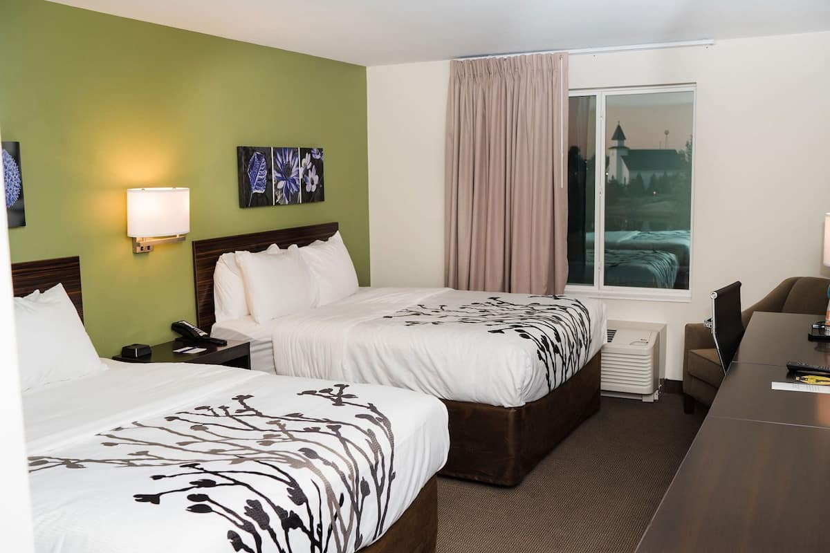 A hotel room with two beds, a desk, a chair, a nightstand, a lamp, a painting, and a window with curtains.