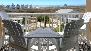 En balkon med to hvide Adirondack-stole og et lille bord, med udsigt over en strand med huse og havet.