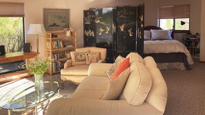 Una sala de estar con un sofá color beige, una mesa de centro de vidrio, una estantería, un mueble negro con obras de arte, una cama con ropa de cama blanca y una ventana con persianas.