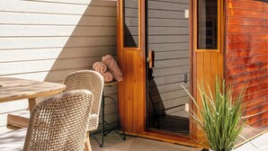 Een houten tuinhuis met een schuifdeur, rieten stoelen, een tafel en een bloempot.