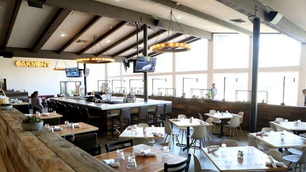 Een restaurant met een lange houten bar, diverse tafels en stoelen, en grote ramen voor veel natuurlijk licht.