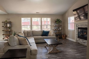 A living room with a sectional sofa, a coffee table, and a fireplace. There are two windows with blinds, and a bookshelf with plants.