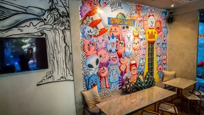 A room with a wall mural featuring colorful cartoon characters and a sign that reads "STELLAR CAPSULES". There is a flat-screen TV mounted on the wall, a wooden table, and a few chairs. - Stellar Capsules - Hostel (Seminyak)