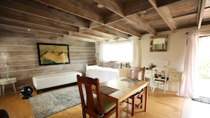 Une pièce chaleureuse avec un plafond en bois, un lit doté d’une tête de lit blanche, une table à manger avec des chaises, un petit bureau et un grand miroir au mur.