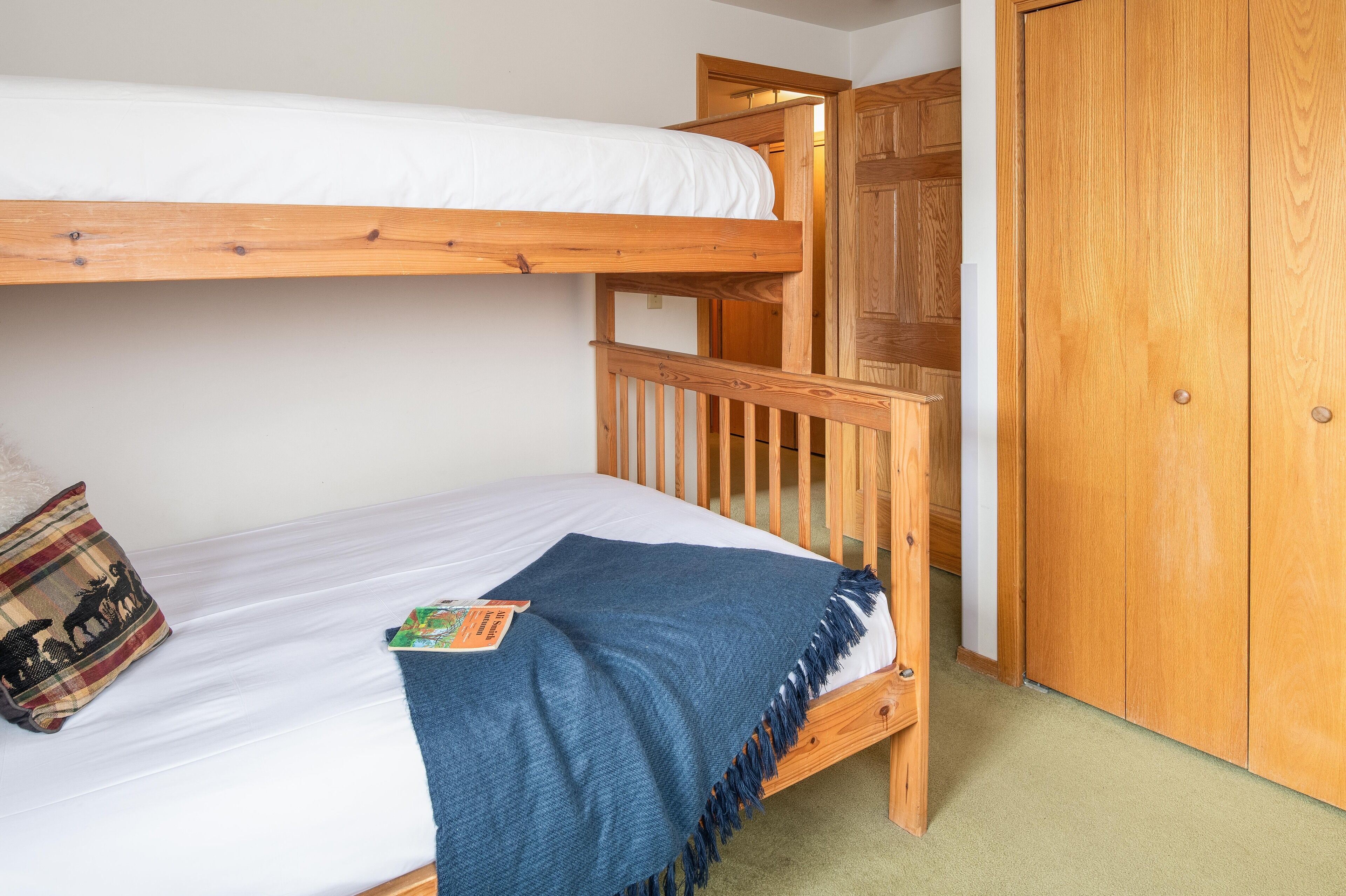A bunk bed with a wooden frame, a white mattress, a blue blanket, and a plaid pillow.