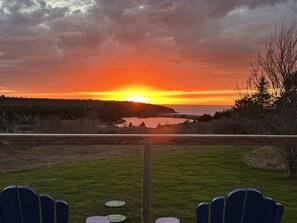 Une vue sur le coucher de soleil depuis une terrasse équipée de chaises et d’une table, donnant sur une étendue d’eau et une zone boisée.