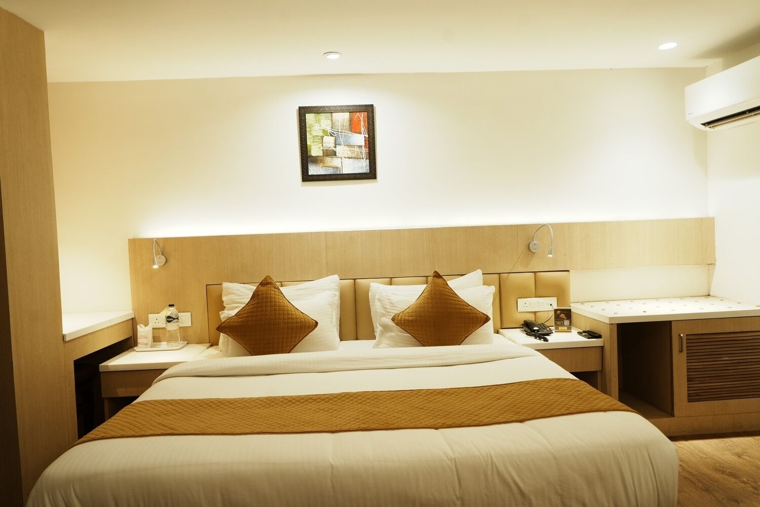A hotel room with a large bed, two bedside tables, a nightstand, a wall-mounted air conditioner, and a framed picture above the bed.