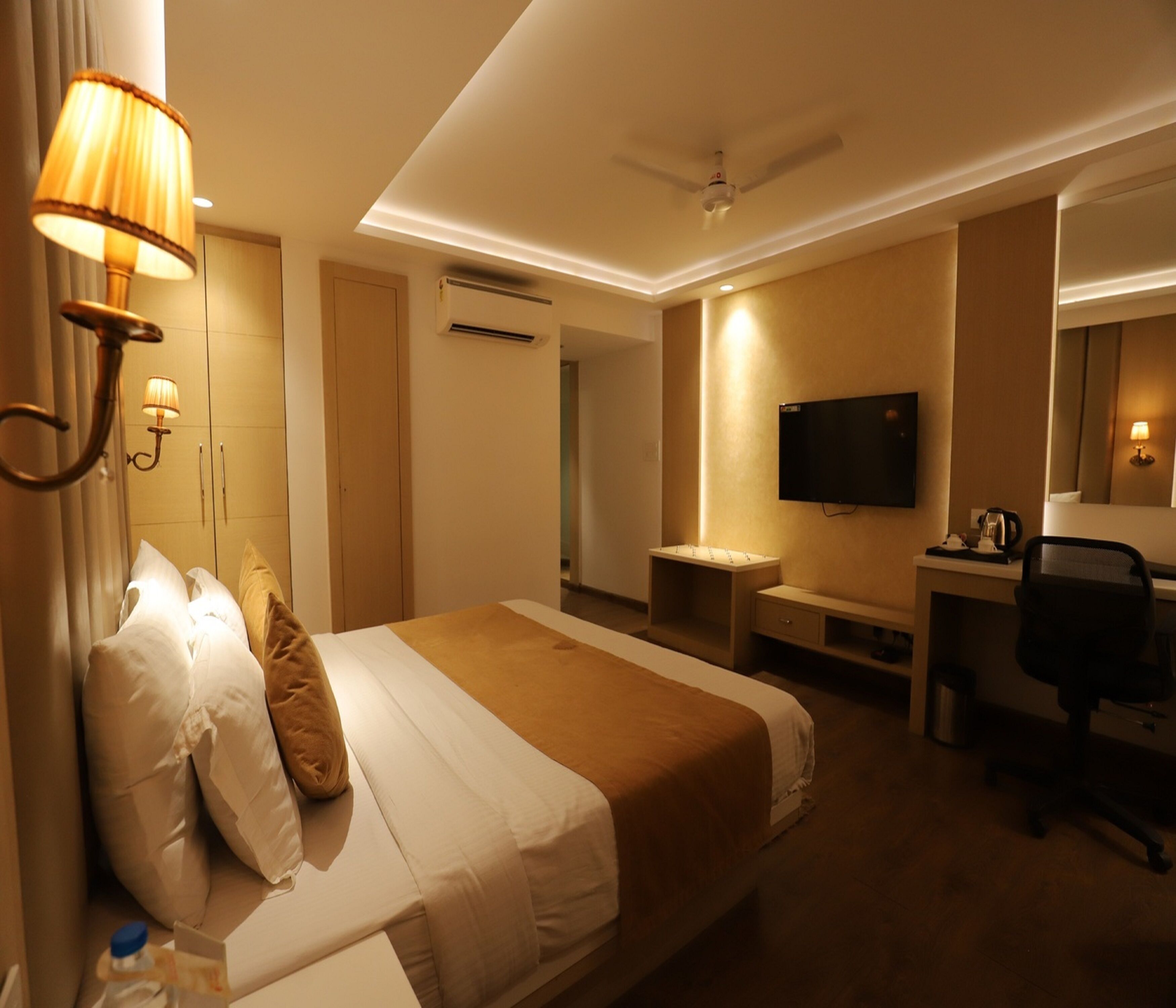A hotel room with a bed, bedside lamp, TV, desk, chair, and a ceiling fan.