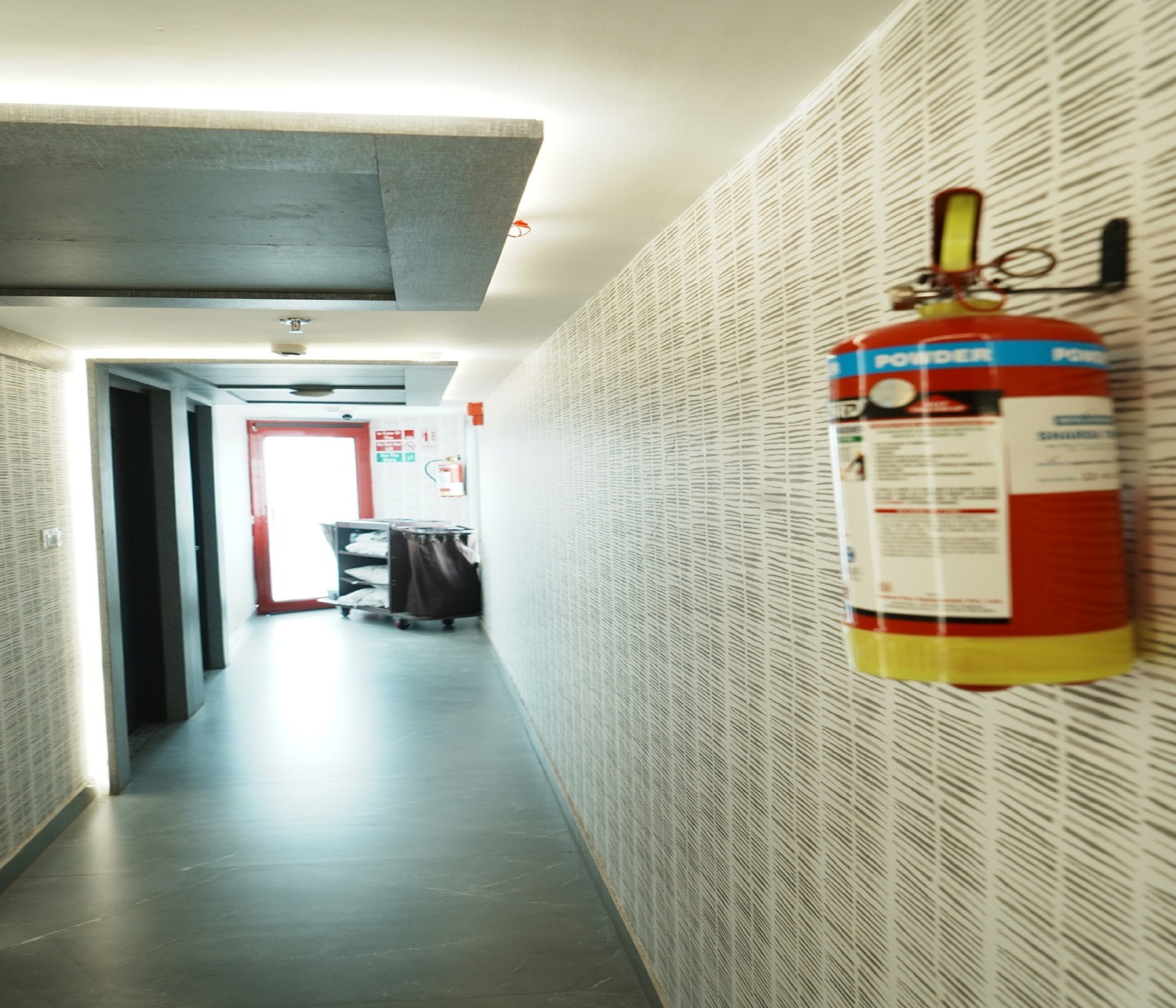 A hallway with a fire extinguisher mounted on the wall, a ceiling vent, and a door at the end.