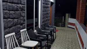 A covered outdoor seating area with chairs and tables, a stone wall, and a tiled floor. - Pear Tree Inn (Kotagiri)