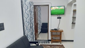 A modern living room with a grey sofa, a flat-screen TV mounted on the wall, a wooden side table, and patterned curtains. - Pear Tree Inn (Kotagiri)