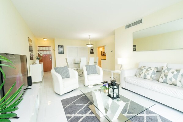 A modern living room with white furniture, a glass coffee table, and a large mirror.