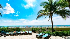A beachside lounge area with sun loungers, a palm tree, and a view of the ocean. - Beachfront Apt | Balcony + Pool | 4 adults + kids (Hollywood)