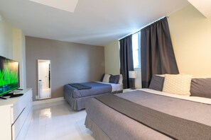 A hotel room with a bed, a desk with a television, a bedside table with a lamp, and a door leading to another room.