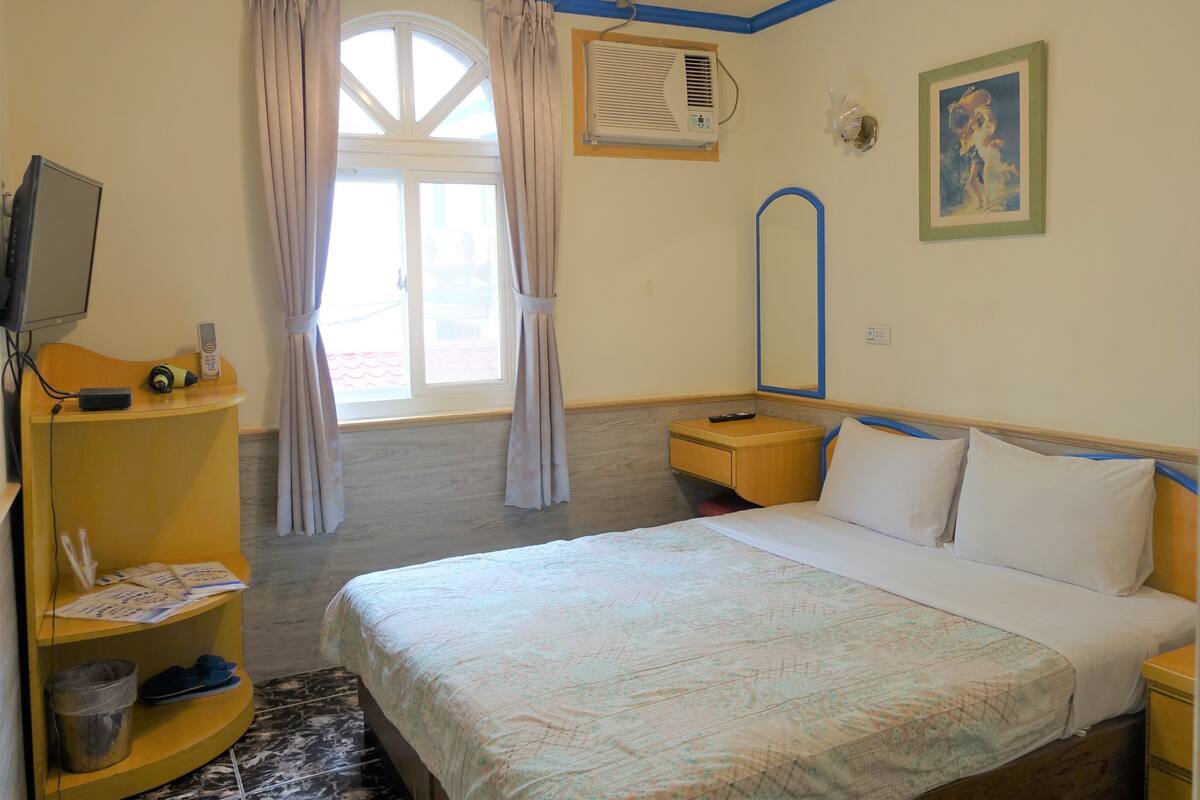 Une chambre d’hôtel comprenant un lit, un bureau avec un ordinateur, une chaise, une fenêtre avec des rideaux, un appareil de climatisation et un tableau accroché au mur.