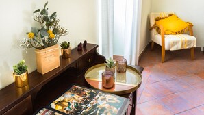 Ein gemütliches Zimmer mit einem Holzboard, einem runden Glastisch mit Kerzen, einem Sessel mit gelbem Kissen und einer Wand mit Pflanzen.