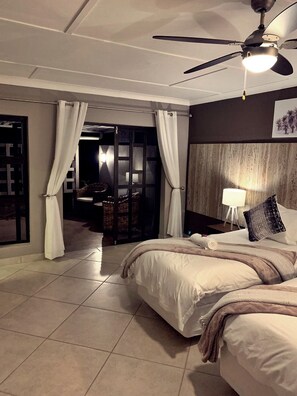 Habitación de hotel con cama, ventilador de techo, mesita de noche con lámpara y vistas a un balcón.