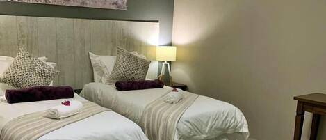 Habitación de hotel con dos camas, una mesita de noche con lámpara, cabecera de madera y un cuadro de árboles en la pared.