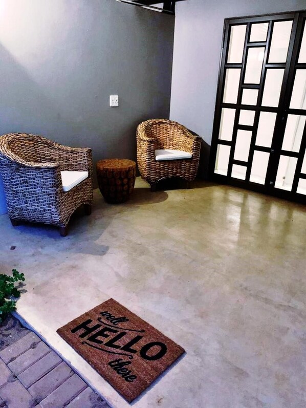 Una habitación con dos sillas de mimbre, un taburete pequeño y redondo, y un felpudo con la inscripción 'HELLO'.