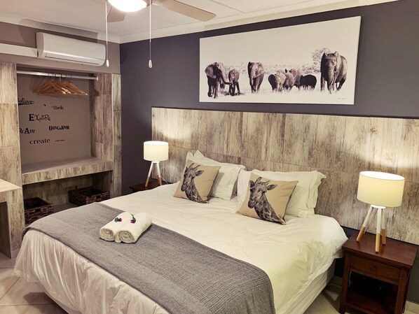 A bedroom with a large bed, two bedside lamps, a wall-mounted air conditioner, and a framed picture of elephants. - The Wilds Guest House (Hluhluwe)
