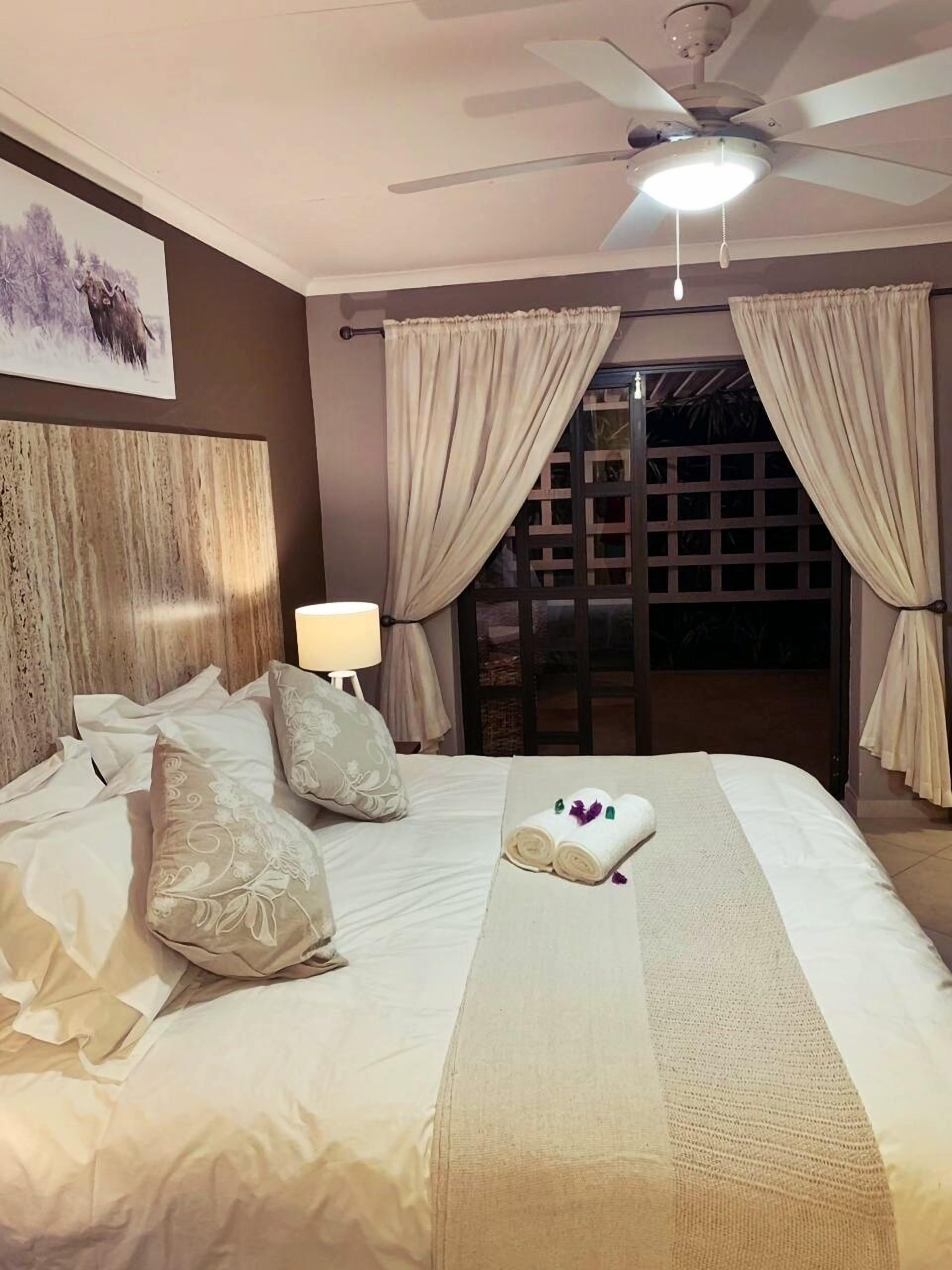 Une chambre d’hôtel avec un lit, des rideaux, un ventilateur de plafond et une vue sur l’extérieur, visible à travers une porte.