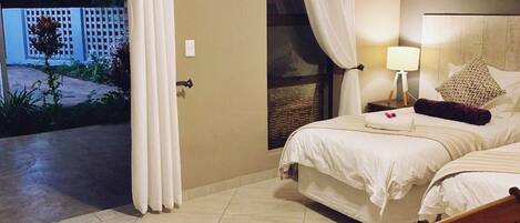 A hotel room with a bed, a ceiling fan, a nightstand, a lamp, and a view of the outside.