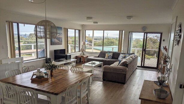 A spacious living area with a wooden dining table, white chairs, a grey sofa, and a TV. Large windows offer views of the outdoors.