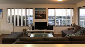 A living room with a flat-screen TV, two sofas, a coffee table, and large windows with blinds.