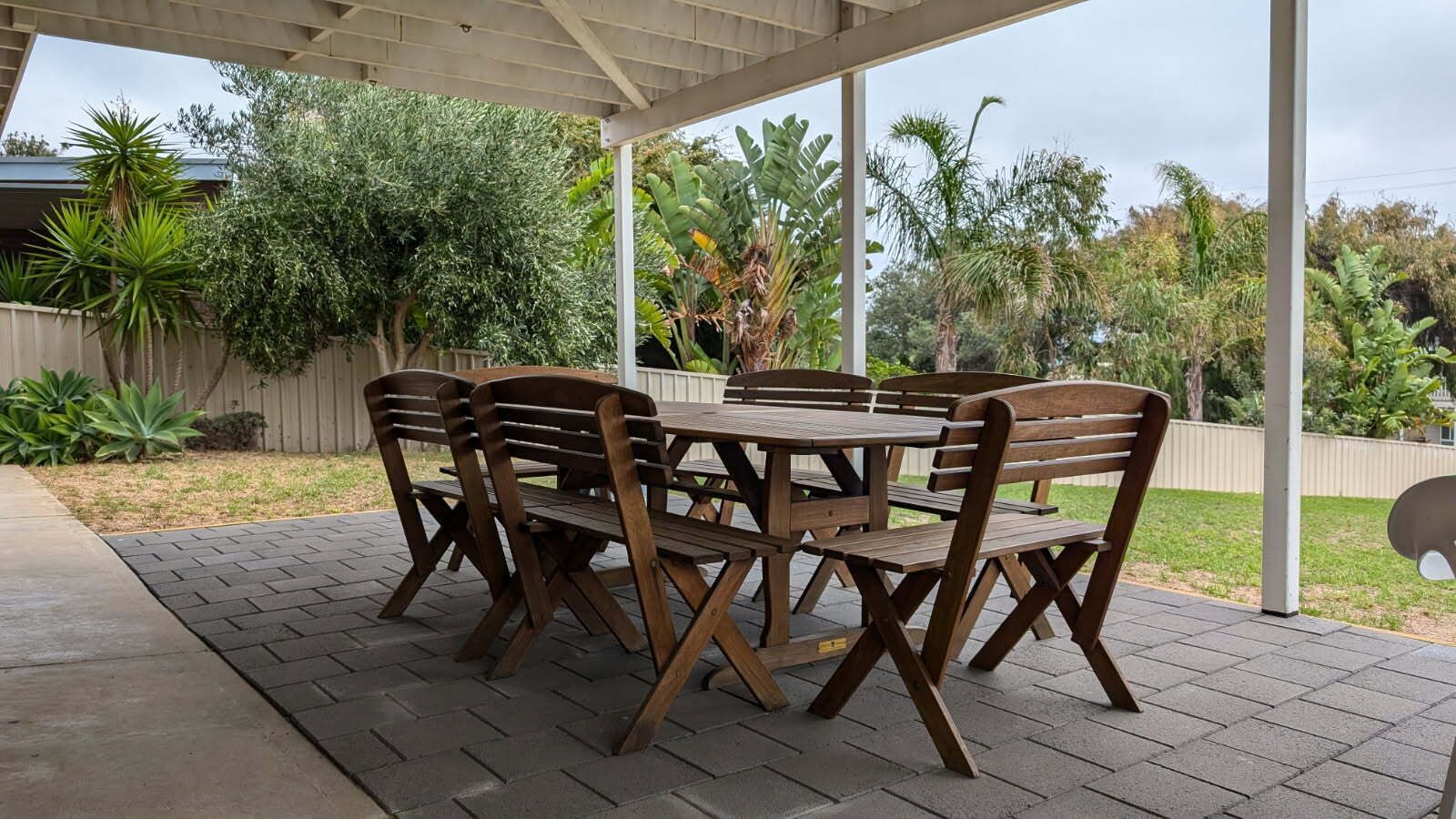 A covered patio with a wooden table and chairs, surrounded by a garden with various plants and a wooden fence.