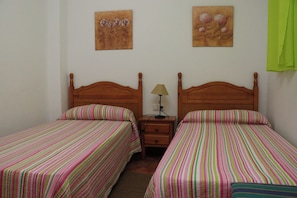 A room with two single beds, wooden headboards, striped bedding, a nightstand with a lamp, and two wall-mounted paintings.