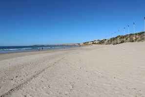 Une vaste plage de sable fin, un ciel bleu dégagé, une mer calme et une falaise lointaine.