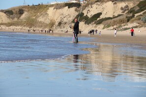 A beach scene with a person fishing, sandy dunes, and people in the background. - Apartment with air-con in lounge (CONIL DE LA FRONTERA)