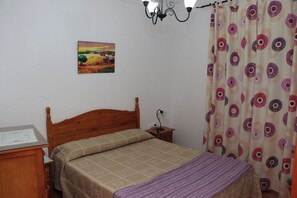 Une chambre avec un lit en bois, une table de chevet, un bureau avec une lampe, un tableau au mur et un rideau à motifs.