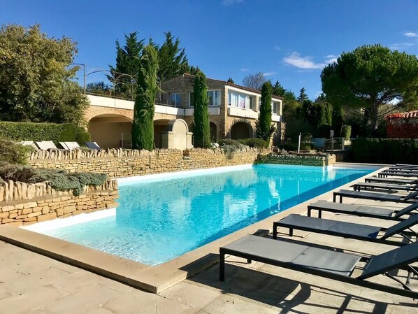 A swimming pool with lounge chairs, a stone wall, and a building with arches in the background.