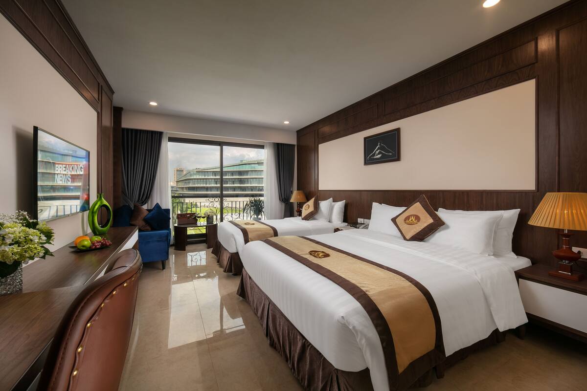 A hotel room with two beds, a desk, a chair, a TV, and a balcony with a view of buildings.