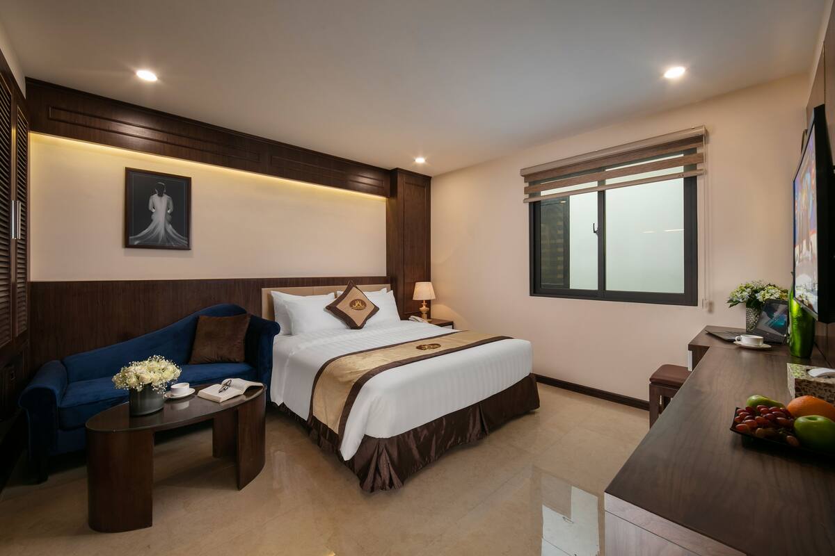 A hotel room with a bed, a blue sofa, a small table with flowers, a window, and a fruit bowl on the nightstand.