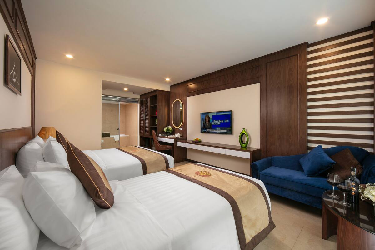 A hotel room with two beds, a blue sofa, a TV, and a small table with a vase and wine glasses.