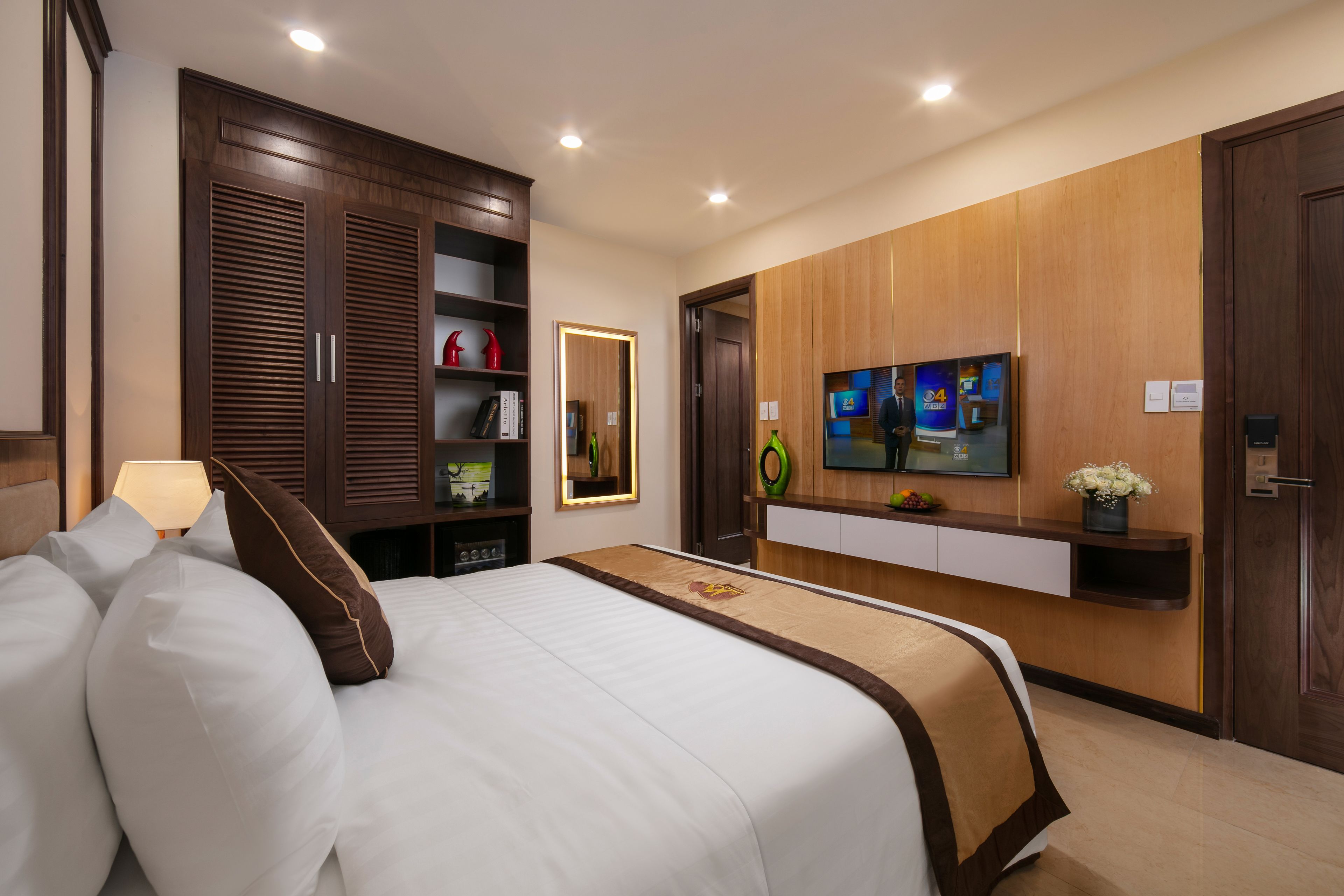 Une chambre d’hôtel avec un grand lit, une armoire en bois, un téléviseur fixé au mur et une petite étagère ornée d’objets décoratifs.