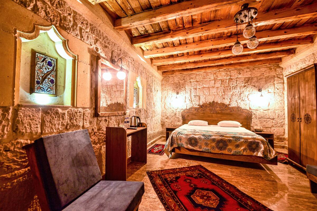 A rustic bedroom with a stone wall, wooden ceiling beams, a bed with a patterned bedspread, a wooden chair, and a red rug.