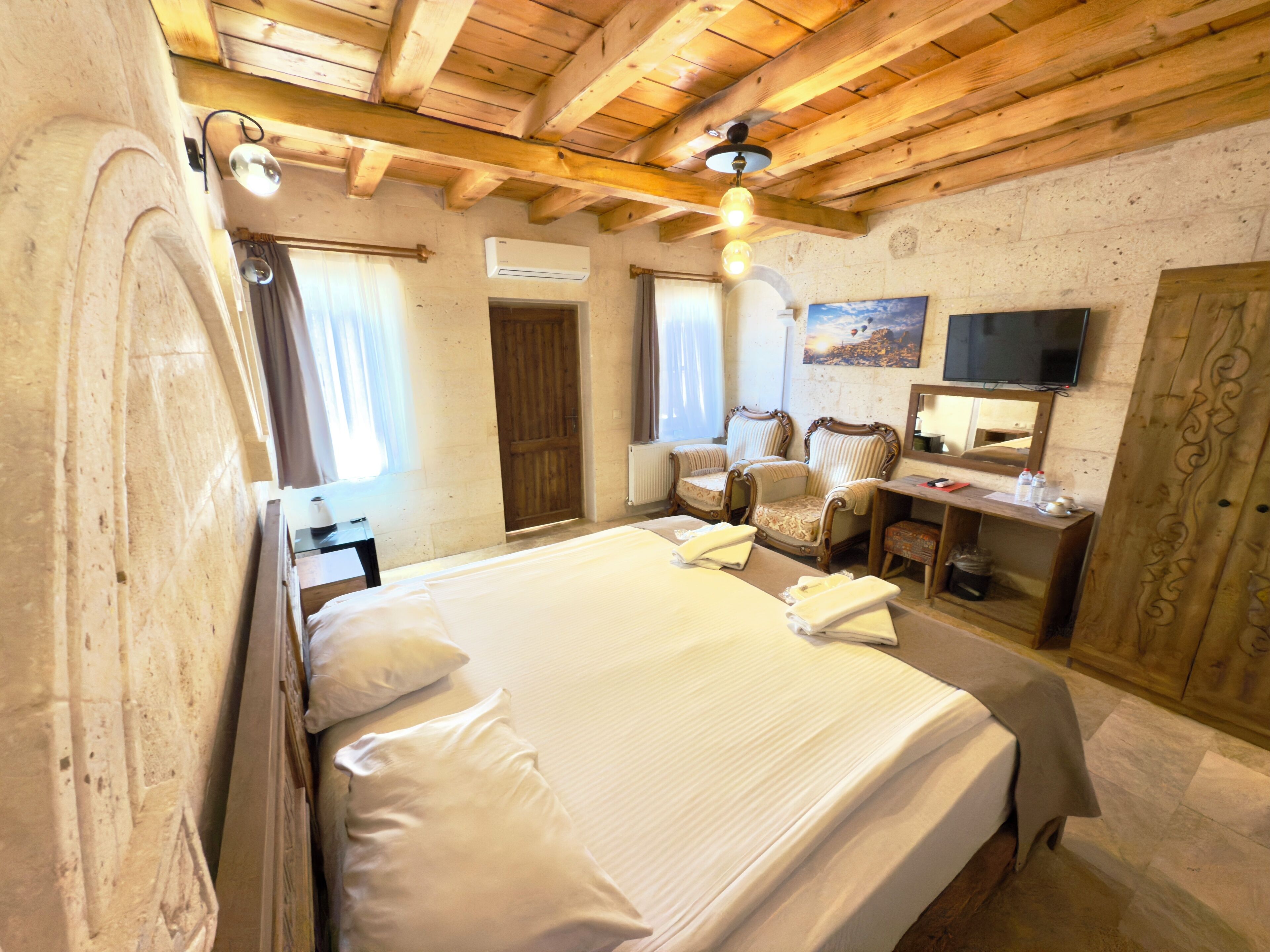 Un dormitorio con una cama grande, vigas de madera en el techo, un escritorio con silla, un televisor y un espejo.