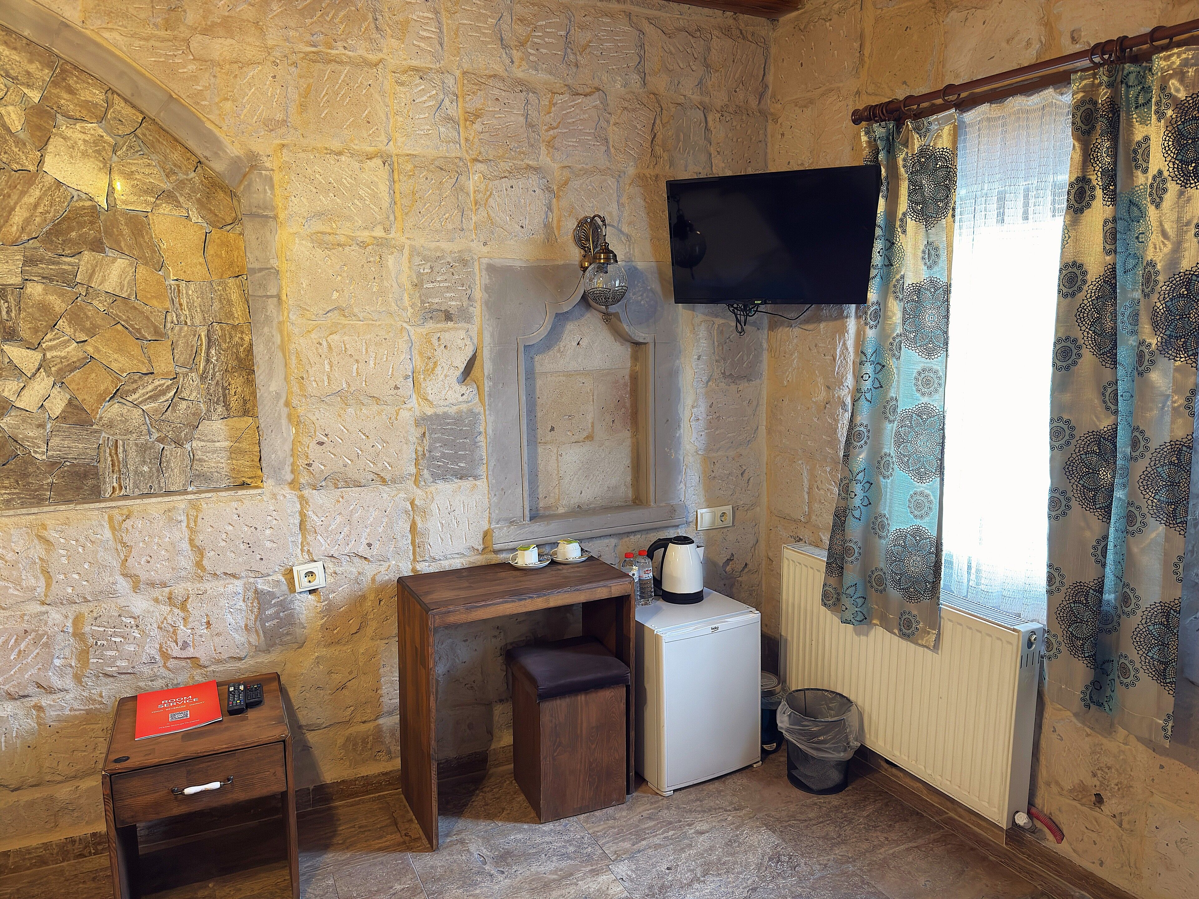 A stone-walled room with a wooden desk, a small refrigerator, a radiator, and a window with patterned curtains.