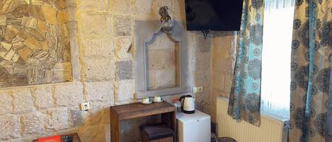 A stone-walled room with a wooden desk, a small refrigerator, a radiator, and a window with patterned curtains.