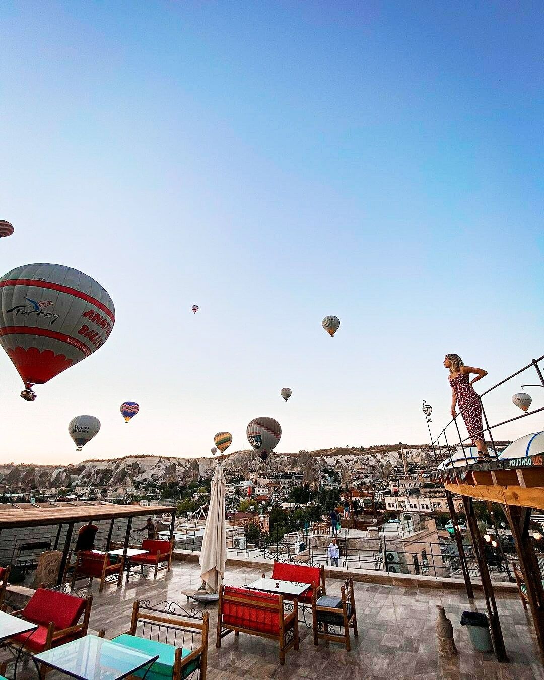 A rooftop terrace with tables and chairs, overlooking a town with buildings and a view of the sky with hot air balloons.