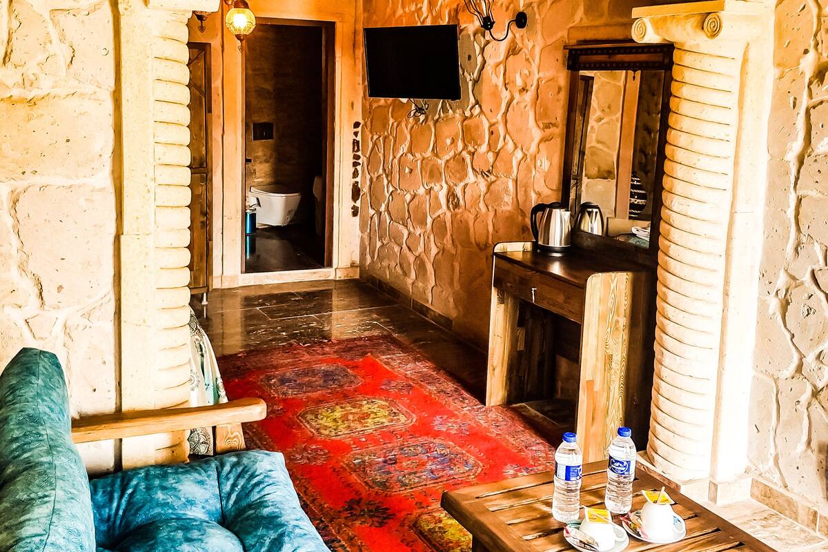A room with a stone wall, a wooden bench, a red patterned rug, a wooden table with a water bottle, and a television mounted on the wall.